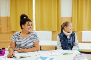 Två unga flickor sitter vid sina pulpeter i skolan, de ler och titta till höger. Bakom dem är en solgul vägg och solgula gardiner.