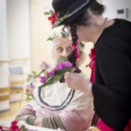 På bilden tittar en äldre dam på då en person utklädd till clown binder en blomsterkrans.