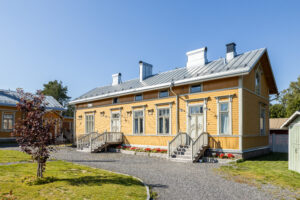 En grusgång leder fram till ett välskött gult trähus. Himlen är sommarblå och gräsmattan är kortklippt och grön.