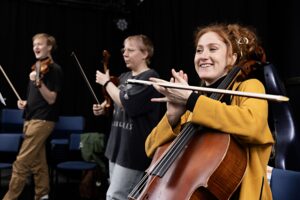 En cellist i gul tröja ler och klappar händerna med stråken mellan fingrarna. I bakgrunden knäpper håller en annan person sin violin som en gitarr och knäpper på den.