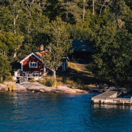Ett fotografi föreställande en strandtomt i skärgården. På fotot ser vi en strandkant, en brygga och en röd stuga.