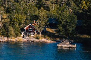 Ett fotografi föreställande en strandtomt i skärgården. På fotot ser vi en strandkant, en brygga och en röd stuga.