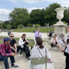 En bild tagen under praktikantträffen i Paris, sensommaren 2023. På bilden ser vi ett gäng unga vuxna som sitter på gröna stolar i en park i Paris.