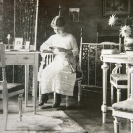 Ett gammalt svartvitt fotografi från 1910- eller 20-talet, föreställande en ung dam som sitter på en stol. Hon är iklädd tidstypisk vit dräkt och sitter med blicken fäst i det handarbete hon har i sina händer.