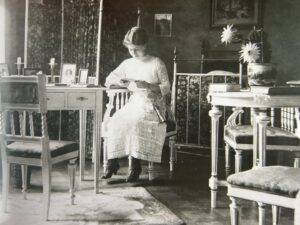 Ett gammalt svartvitt fotografi från 1910- eller 20-talet, föreställande en ung dam som sitter på en stol. Hon är iklädd tidstypisk vit dräkt och sitter med blicken fäst i det handarbete hon har i sina händer.