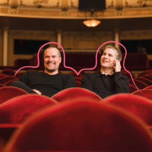Två män sitter i en teatersal, tittar mot scenen och skrattar.