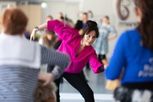 En kvinna i mörk hår och en grannrosa skjorta dansar i en dansstudio.