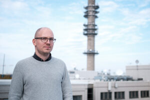 En skallig man i mörka glasögonbågar tittar mot kameran. I bakgrunden syns Yles tv-torn.