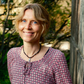 På bilden syns Sophie Henricson. Hon har blont vågigt hår, en blus i rött.blått och vitt. Hon ser in i kameran och ser glad ut.