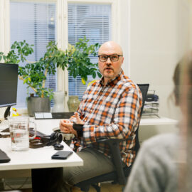 På bilden syns en person som sitter vid sitt arbetsbord. Han har rutig skjorta och glasögon med mörka bågar. Han är vänd mot kameran och talar med en person som vi bara ser ryggen av.