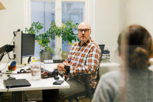 På bilden syns en person som sitter vid sitt arbetsbord. Han har rutig skjorta och glasögon med mörka bågar. Han är vänd mot kameran och talar med en person som vi bara ser ryggen av.
