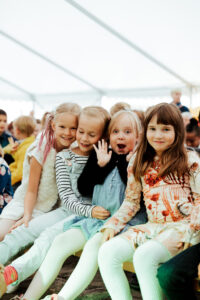 På bilden syns fyra flickor. De sitter på en bänk i ett festtält och alla ser glada och spralliga ut. Flickan i mitten vinkar till fotografen.