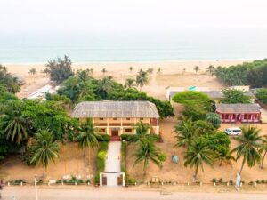 Bild föreställande Villa Karo i Grand-Popo, Benin. Ett tvåvåningshus invid en strand omgiven av palmer.