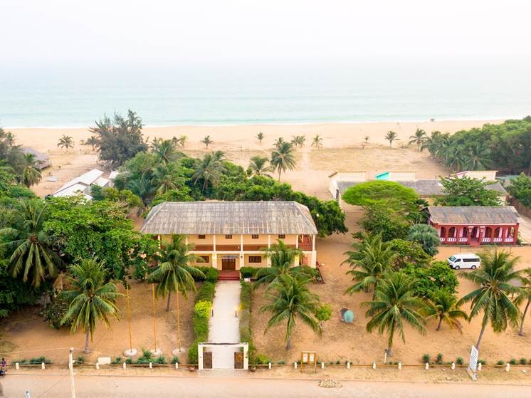 Bild föreställande Villa Karo i Grand-Popo, Benin. Ett tvåvåningshus invid en strand omgiven av palmer.