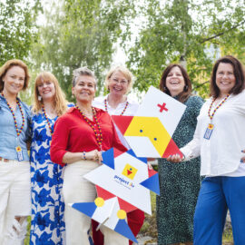 Sex damer i färgglada kläder står på rad och tittar in i kameran. Alla ser glada ut. Bakom dem syns några gröna trädkronor.