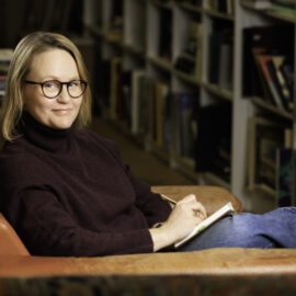 En kvinna iklädd en plommonröd polotröja, jeans och sneakres sitter i en läderfotölj med ett anteckningsblock i knät. Hon har glasögon och blpond acellångt hår och tittar glatt in i kameran