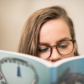 En person med mörkbågade glasögon och mörkt hår läser en bok. Munnen och hakan skyms av boken.