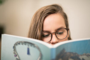 En person med mörkbågade glasögon och mörkt hår läser en bok. Munnen och hakan skyms av boken.