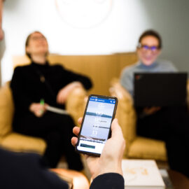 En hand håller i en mobiltelefon där Kulturofondens webbplats synd. I bakgrunden sitter två suddiga personer som skrattar gott.