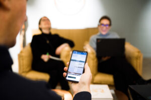 En hand håller i en mobiltelefon där Kulturofondens webbplats synd. I bakgrunden sitter två suddiga personer som skrattar gott.