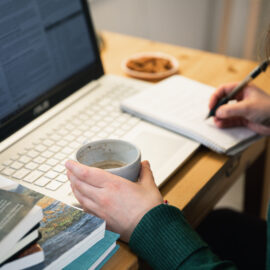 På bilden syns en persons händer som vilar vid ett tangentbord. Den ena handen håller en mugg kaffe och den andra en penna som vilar mot ett block.