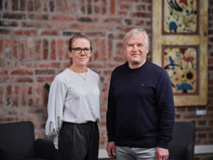 På bilden står Linda Mannila och Stefan Kula framför en röd tegelvägg. Linda har vit blus och Stefan har en blå tröja. De tittar in i kameran och ser tillfreds ut.