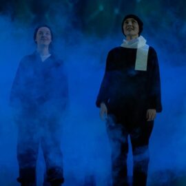 På bilden står två åpersoner i svarta kläder i ett rökmoln. De har vita halsdukar av toalettpappet och svarta mössor. De ser rakt framåt,