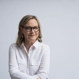 På bilden syns Eva Biaudet. Hon har vit blus, axellångt hår och glasögon med mörka bågar. Hon småler och tittar in kameran.