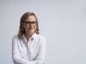På bilden syns Eva Biaudet. Hon har vit blus, axellångt hår och glasögon med mörka bågar. Hon småler och tittar in kameran.