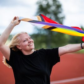 En kvinna står på en springbana och håller en HBTQI+-flagga över huvudet.