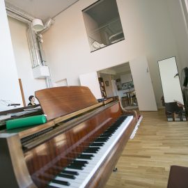 På bilden syns ett stort utrymme med högt i tak. En flygel i förgrunden och en helfigurspegel strax bakom denn. I bakgrunden skymtar ett kök och en fönstervägg till ett rum på andra vångingen. Väggarna är vita och på golvet finns en ljus parkett.